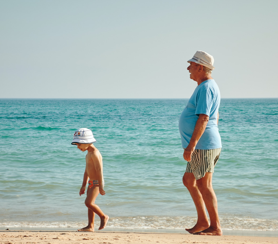 A grandfather strolling along a beach front with his grandson. The sea is blue and the weather is sunny.