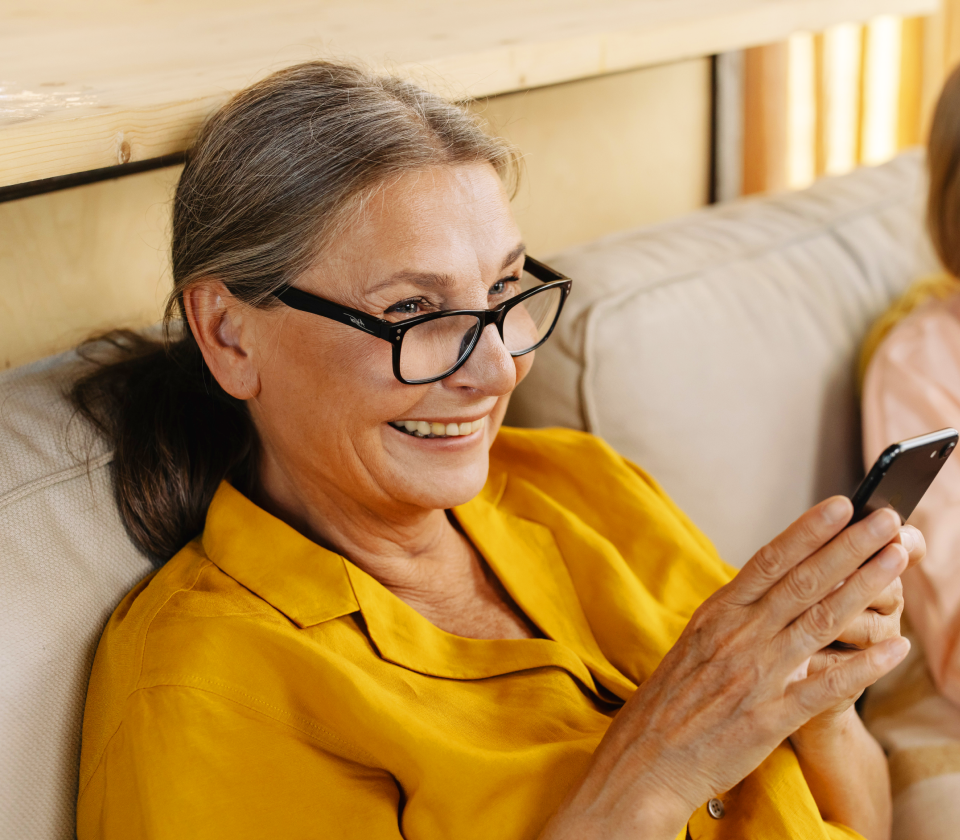 Lady in a yellow shirt wearing glasses and holding and looking at a mobile phone sat on a chair and smiling.