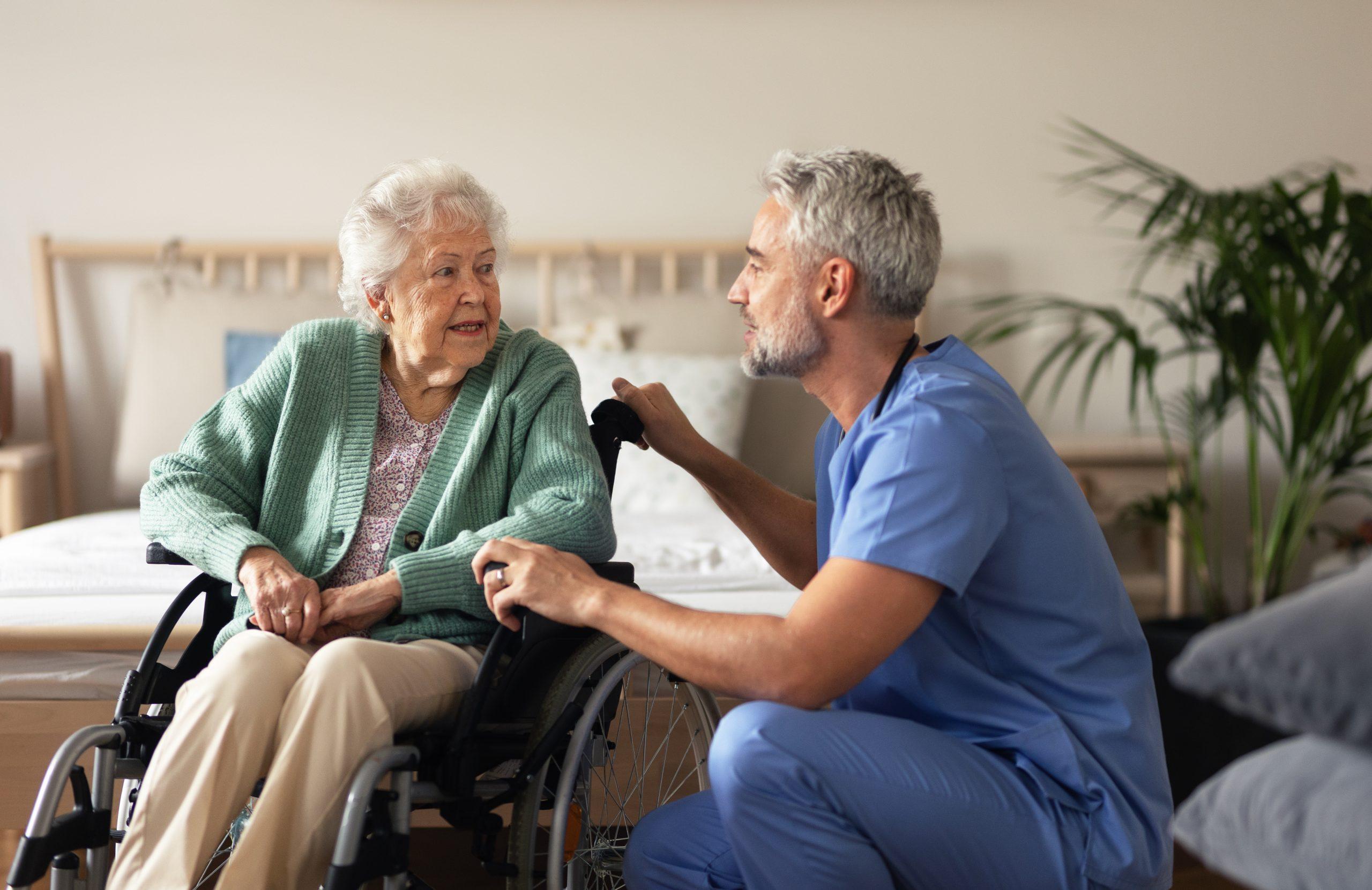 An elderly lady is sat in a wheelchair talking to a male nurse