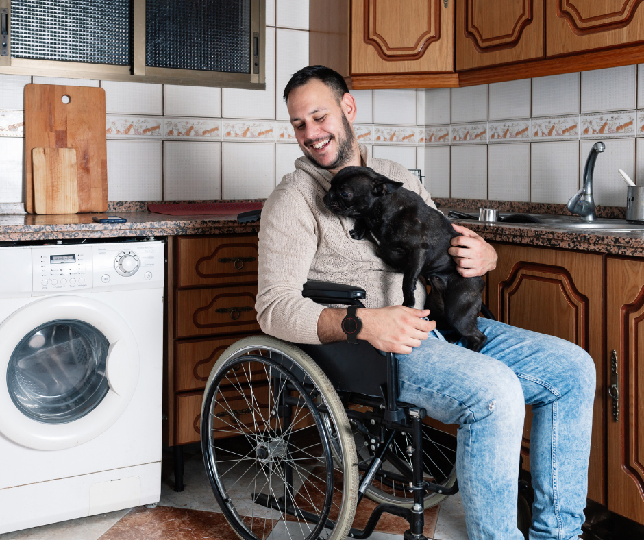 A smiling young man sat in a wheelchair holding onto a black dog on his knees. He is sat in a kitchen and wearing a Yorbl Amica Gero Personal Alarm.